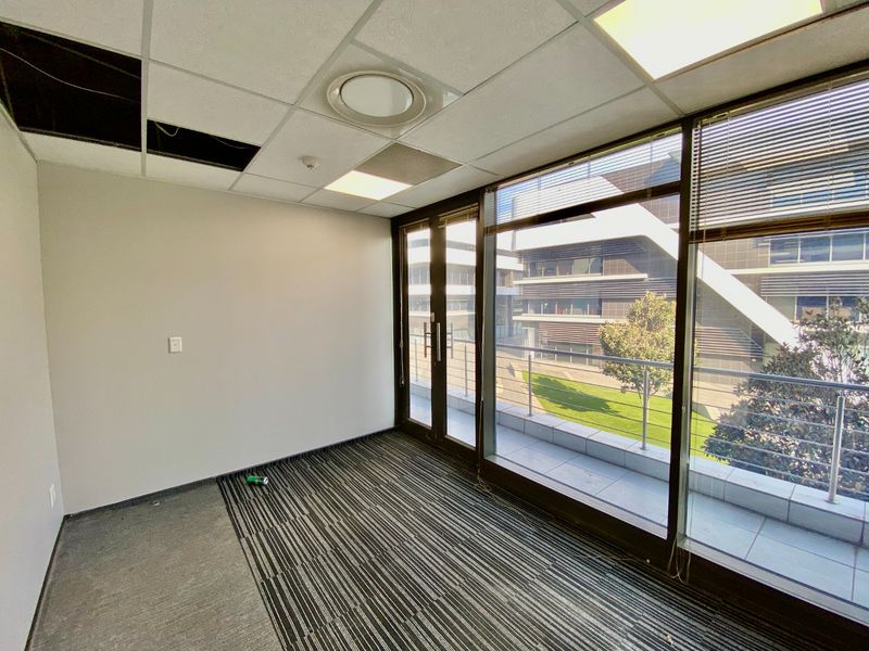 Empty office space featuring a suspended ceiling, floor-to-ceiling windows, and direct access to a private balcony with modern building views.