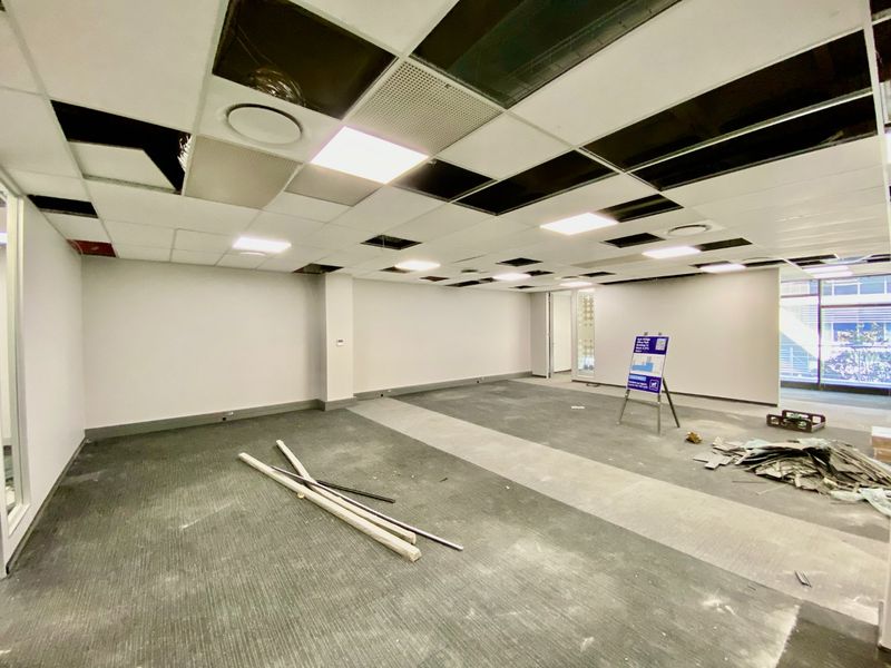 An empty commercial office space featuring a suspended ceiling with missing tiles, recessed lighting, and carpeted flooring, currently undergoing maintenance or renovation.