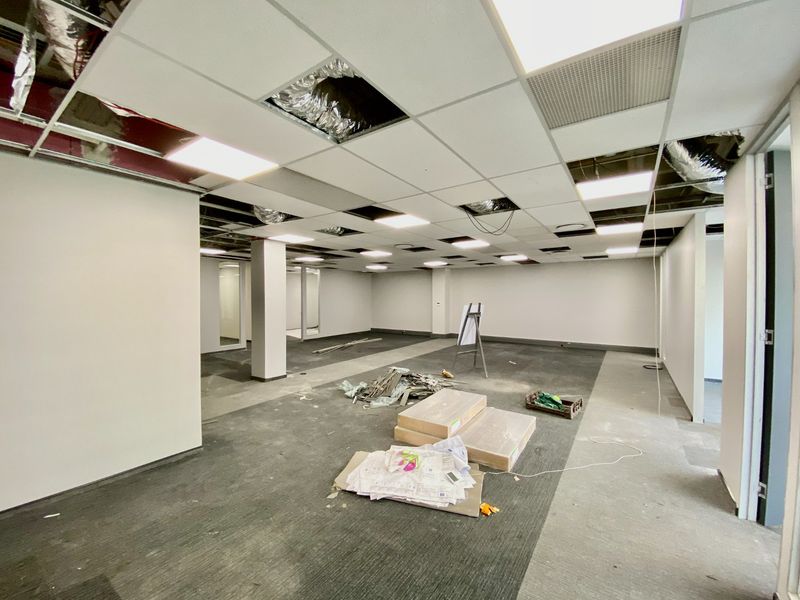 An open-plan commercial office space currently undergoing renovation, featuring a suspended ceiling with missing tiles and exposed infrastructure.