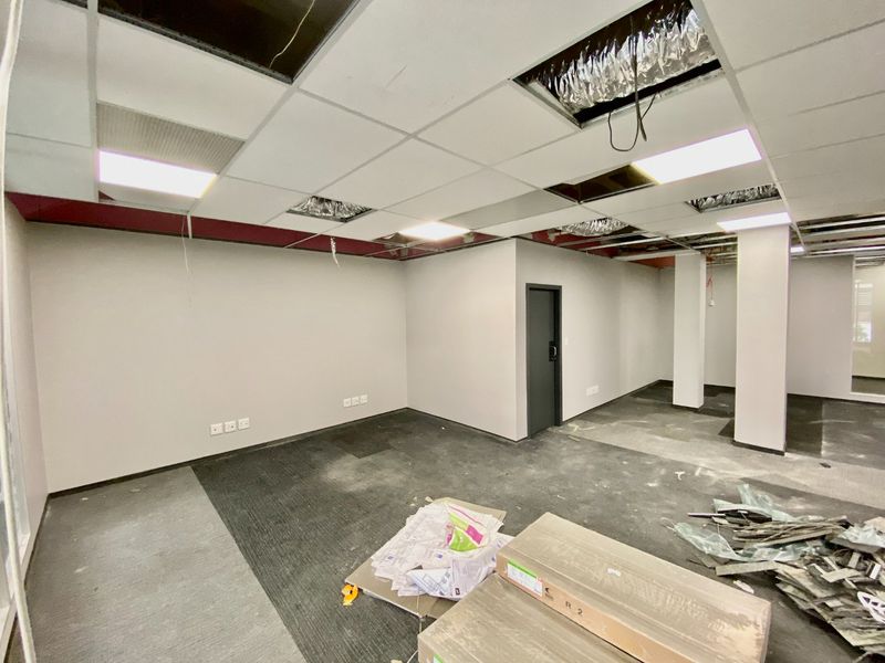 An empty commercial office space featuring a suspended ceiling with missing tiles, carpeted flooring, and structural columns, currently undergoing renovation.