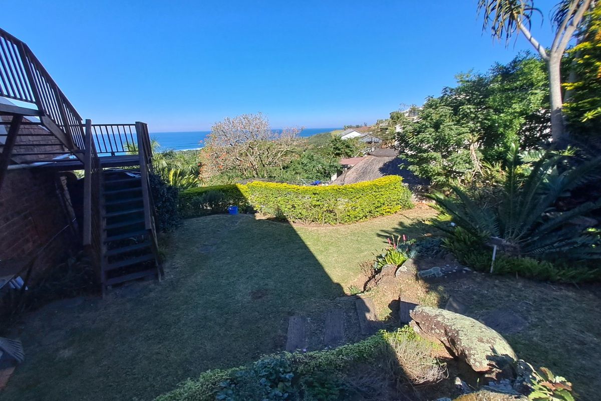 A rear garden area featuring a wooden staircase leading to an upper balcony, with established landscaping and a scenic ocean view.