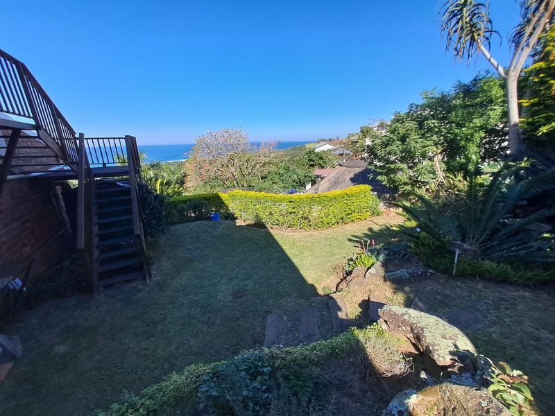 A rear garden area featuring a wooden staircase leading to an upper balcony, with established landscaping and a scenic ocean view.