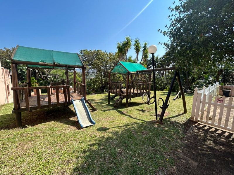 A spacious garden area featuring established trees, a paved walkway, a white picket fence, and wooden play structures with swings.