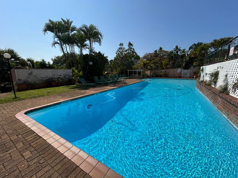 A large, well-maintained swimming pool area featuring brick paving, perimeter walls with lattice fencing, and established garden landscaping.