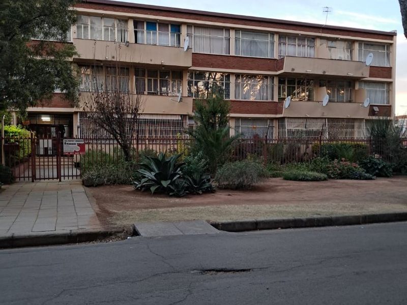 Front elevation of a multi-story apartment building featuring brick facade, private balconies, and secure perimeter fencing with a paved entrance walkway.