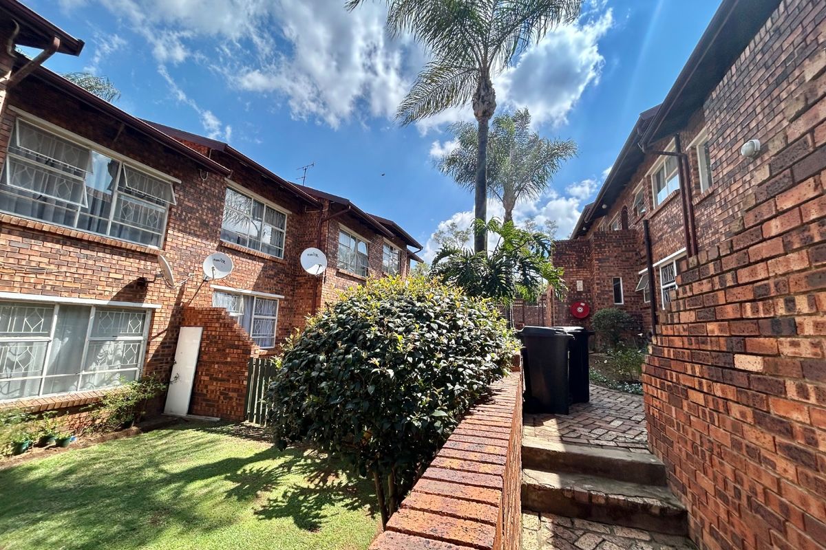 Rear exterior view of a face brick townhouse complex featuring a paved walkway, small lawn area, and established landscaping.