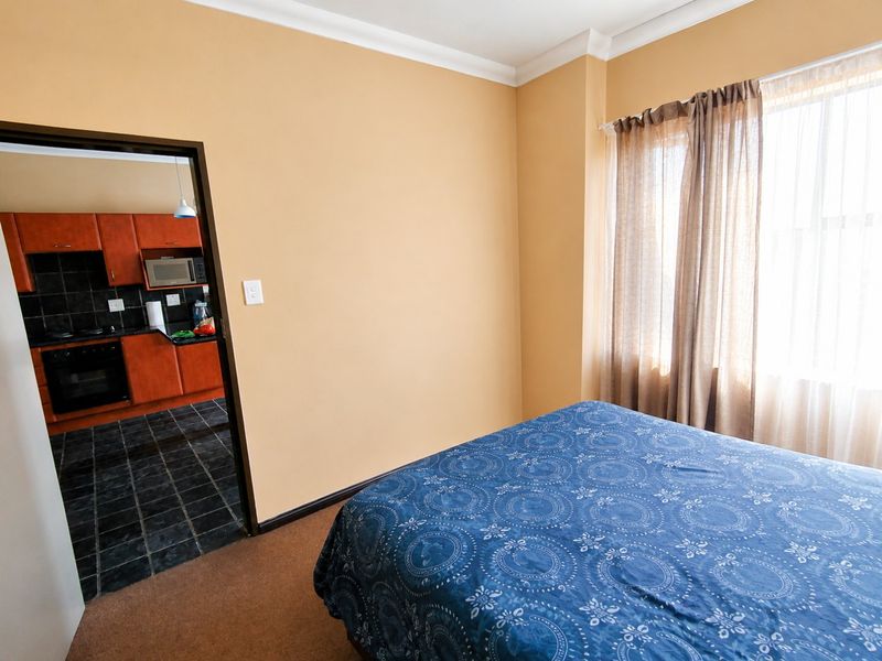 A bedroom with carpeted flooring and a view into an adjacent kitchen featuring dark tiled floors and wood-finish cabinetry.