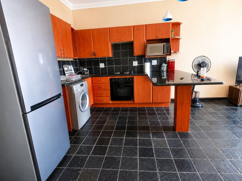 A functional kitchen featuring warm-toned wooden cabinetry, black granite countertops, a built-in oven and hob, and dark tiled flooring with an integrated breakfast bar.