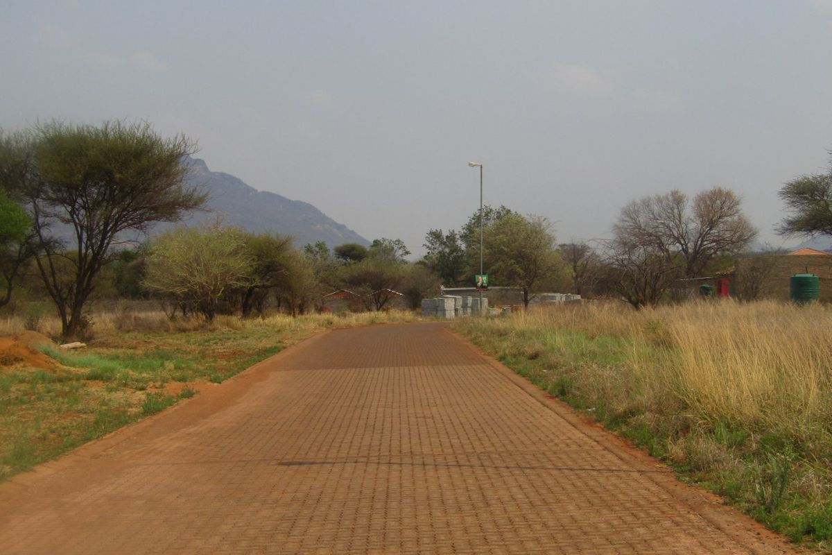 A view of a paved residential access road leading through a natural landscape with surrounding trees and distant mountain views.