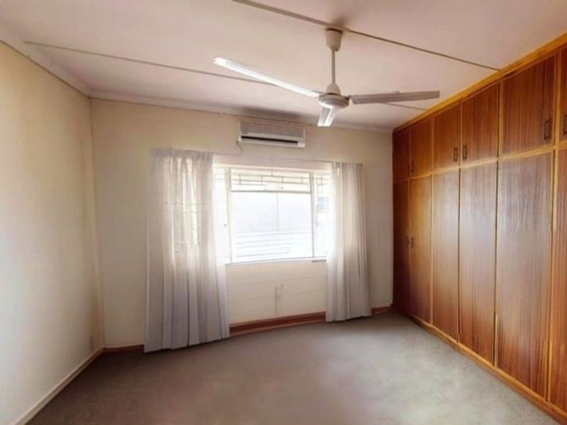 A spacious bedroom featuring extensive built-in wooden cupboards, a ceiling fan, and an air conditioning unit, providing a functional and neutral living space.