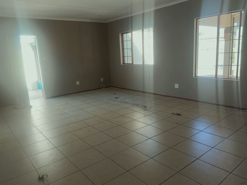 A spacious, empty living area featuring neutral grey walls, tiled flooring, and large windows allowing for natural light.