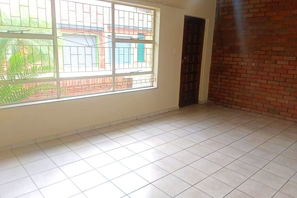 An empty living room featuring tiled flooring, an exposed brick feature wall, and a large window with security bars.