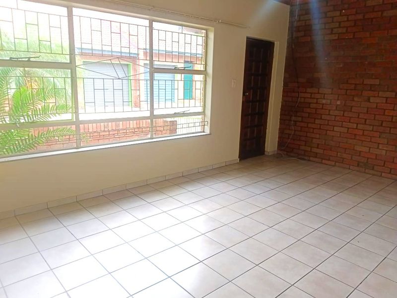 An empty living room featuring tiled flooring, an exposed brick feature wall, and a large window with security bars.