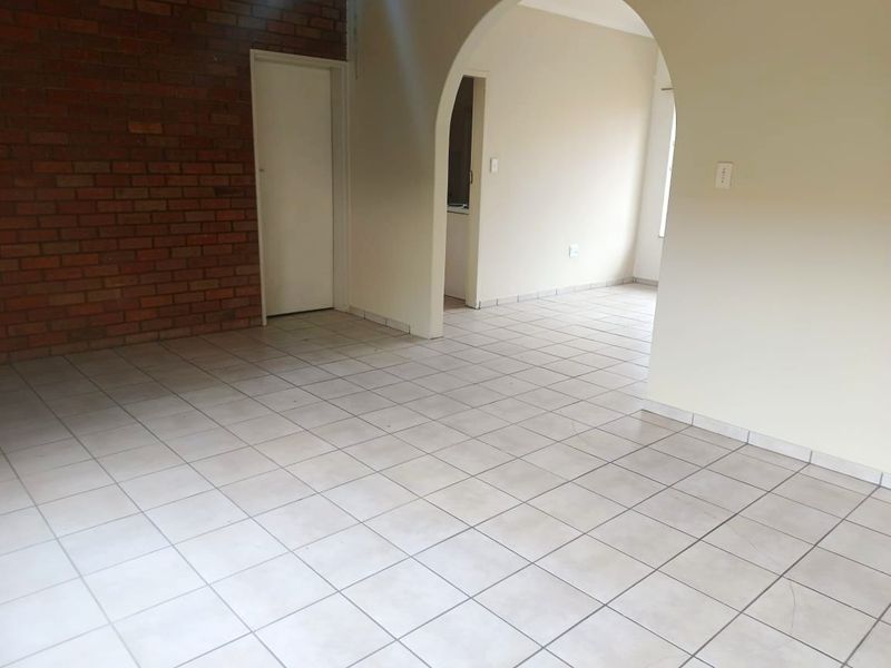 An empty open-plan living area featuring tiled flooring, an exposed brick feature wall, and a classic arched doorway leading to adjacent spaces.