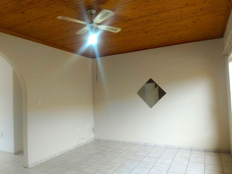 An empty living room featuring ceramic floor tiles, a timber tongue and groove ceiling with a ceiling fan, and a decorative diamond-shaped wall mirror.