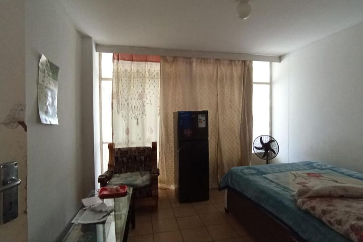 A bedroom featuring tiled flooring and a large window, showing signs of wear including damaged wall plaster near the entrance.