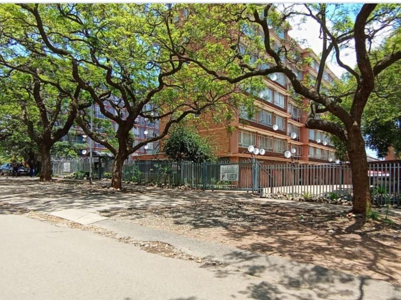Street view of the apartment complex featuring mature trees, perimeter security fencing, and gated access.