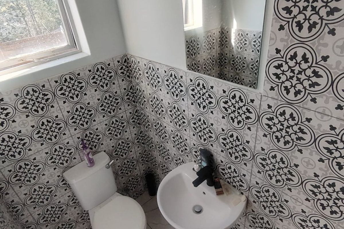 A compact bathroom featuring patterned wall tiles, a pedestal basin, and a toilet, finished with neutral floor tiles and a window for natural light.