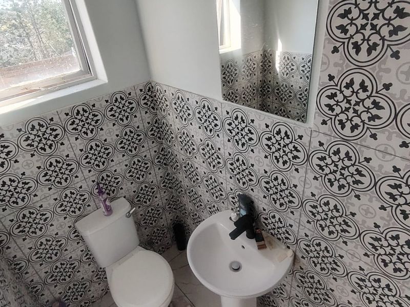 A compact bathroom featuring patterned wall tiles, a pedestal basin, and a toilet, finished with neutral floor tiles and a window for natural light.