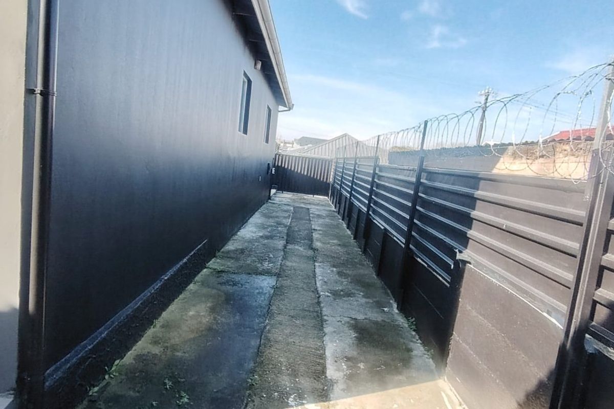 A narrow side passage featuring a concrete walkway, dark painted exterior wall, and metal perimeter fencing topped with security razor wire.