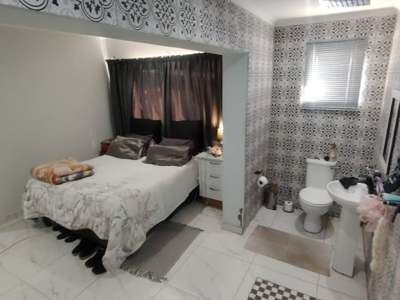 A bedroom featuring an open-plan en-suite bathroom with a pedestal basin, toilet, and rain shower, finished with decorative patterned wall tiles and polished floor tiles.