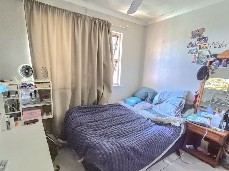 A bedroom featuring tiled flooring, a ceiling fan, and a window with security bars, currently showing signs of wear on the walls and ceiling.