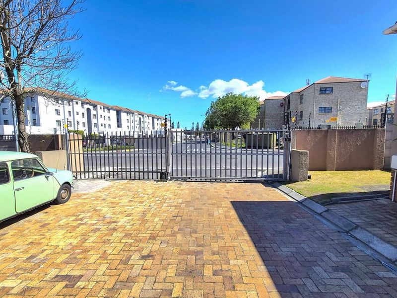 View of the apartment complex entrance featuring a paved driveway, secure metal gate, and perimeter walling with electric fencing.
