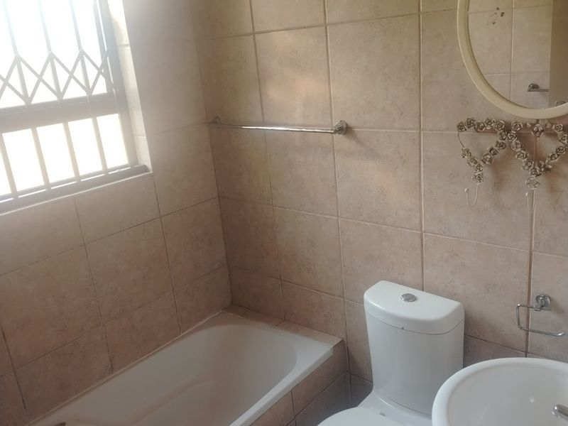 A functional bathroom featuring a bathtub, pedestal basin, and toilet with neutral wall tiling and a window fitted with a security gate.