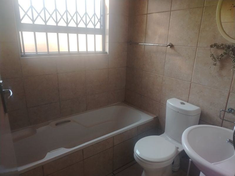 A functional bathroom featuring a built-in bathtub, pedestal basin, and toilet, finished with neutral wall tiling and a security-fitted window.