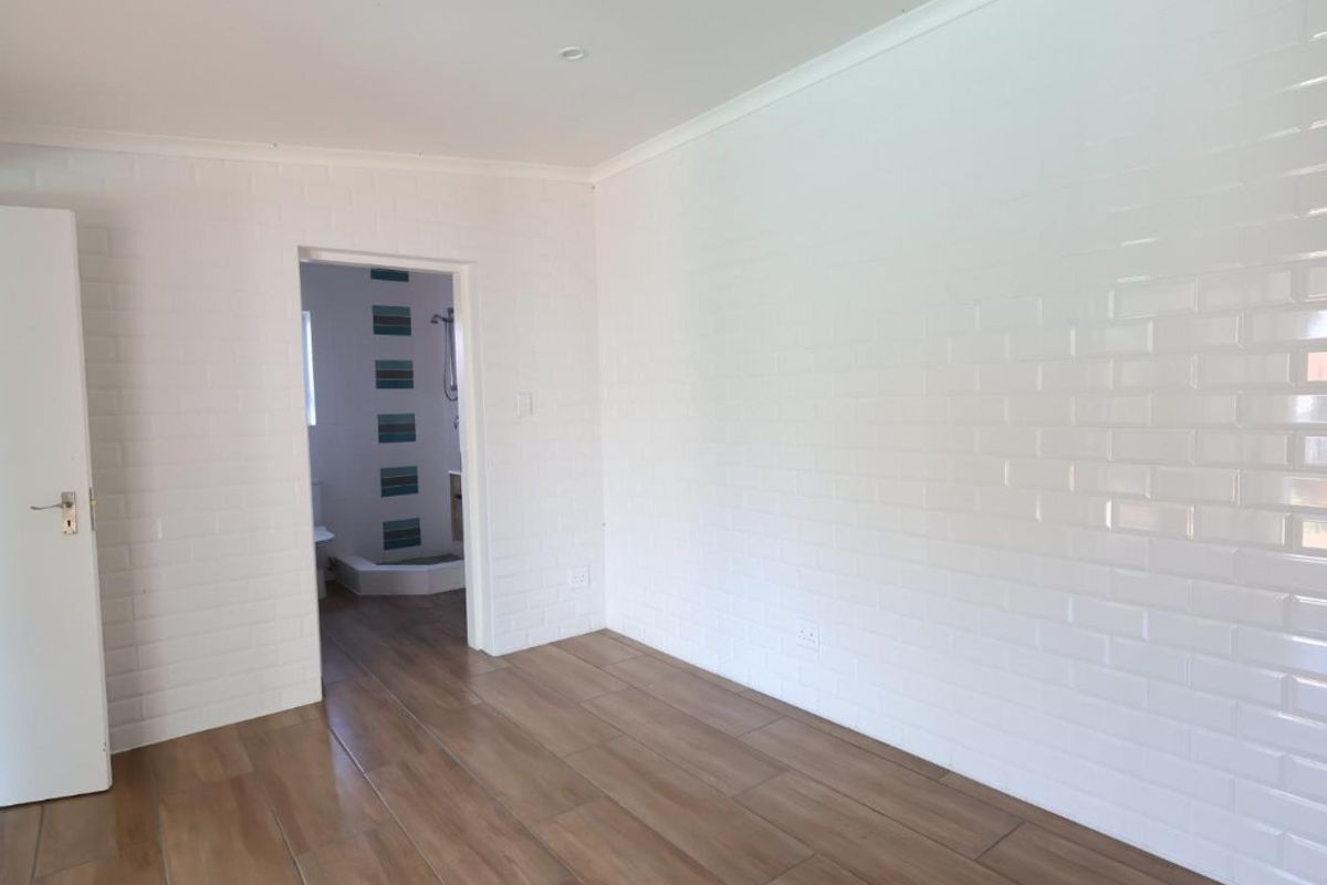 A bright, empty bedroom featuring modern wood-look floor tiles, white subway tile wall cladding, and recessed lighting, with an adjoining bathroom visible.