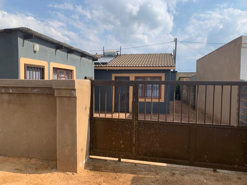 Front view of a residential property featuring a paved driveway, secure perimeter wall, and a house equipped with a solar water heating system.