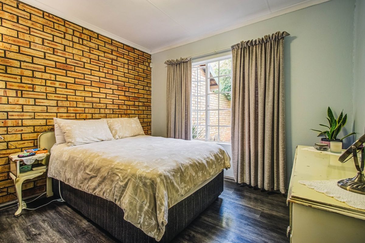 A cozy bedroom featuring a rustic exposed brick feature wall, dark laminate flooring, and natural light through a window.