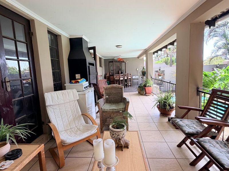 A spacious covered patio featuring tiled flooring, a built-in braai, and a metal balustrade overlooking the garden and pool area.