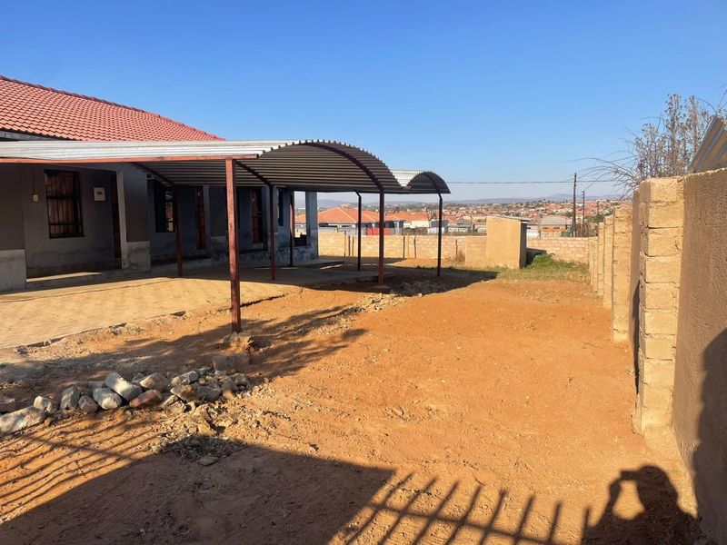 Rear exterior area featuring a corrugated metal carport, paved section, and an expansive unlandscaped yard enclosed by a perimeter wall.