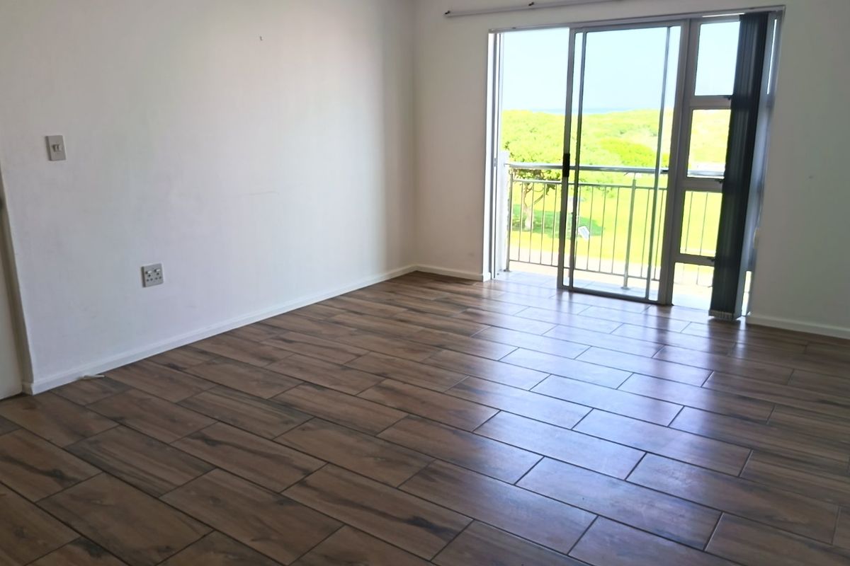 An empty, well-lit living area featuring modern wood-look tile flooring and a sliding glass door that opens onto a balcony with scenic views.