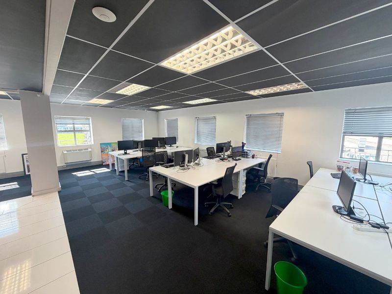 A modern open-plan office space featuring dark carpet tiles, a suspended ceiling with recessed lighting, and multiple windows with venetian blinds.