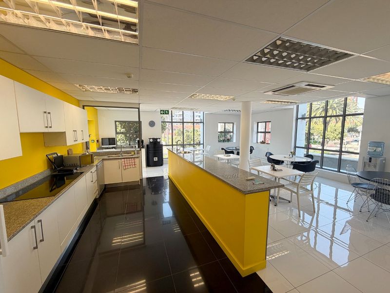 Spacious open-plan commercial kitchen and break area featuring modern cabinetry, granite countertops, high-gloss tiled flooring, and abundant natural light from large windows.
