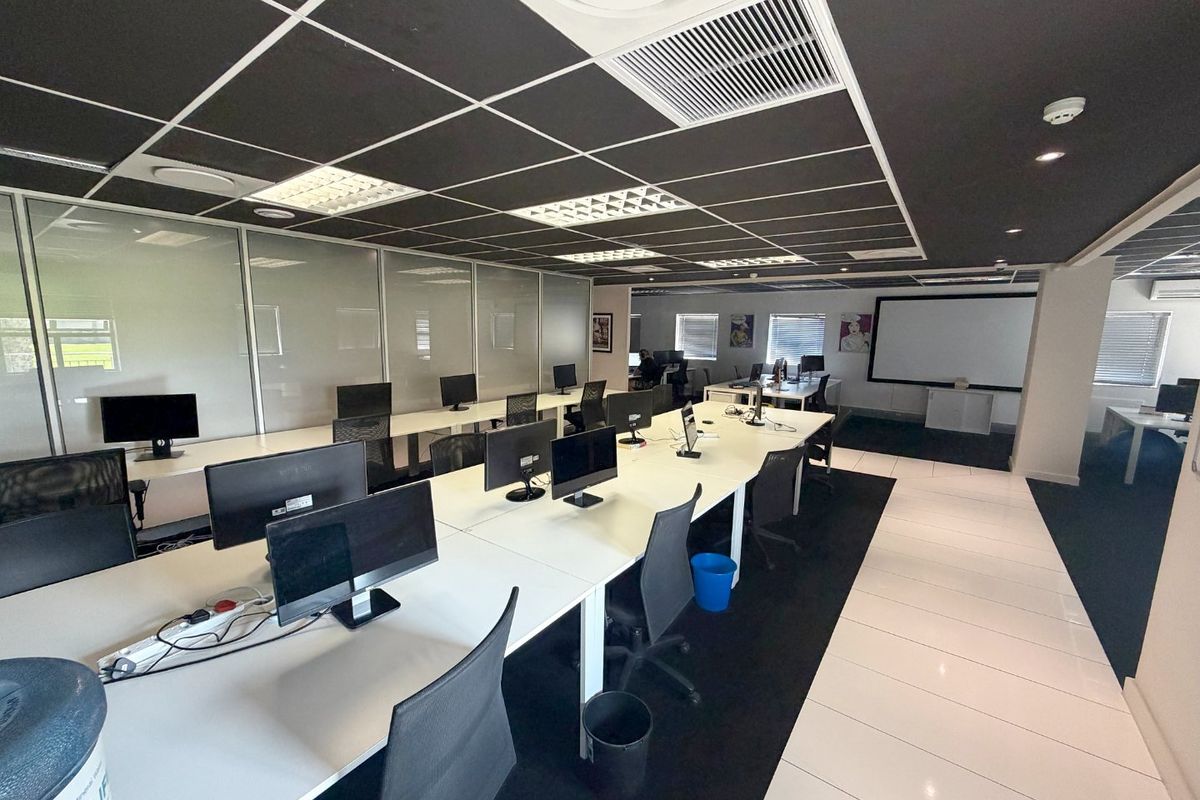 Modern open-plan office space featuring a suspended ceiling, recessed lighting, glass partitions, and a combination of tiled and carpeted flooring with a projector screen.