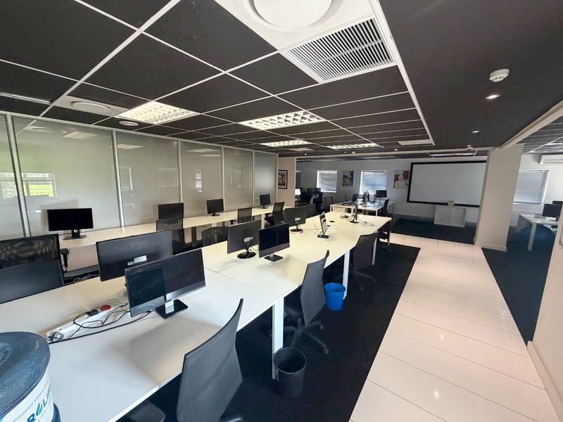 Modern open-plan office space featuring a suspended ceiling, recessed lighting, glass partitions, and a combination of tiled and carpeted flooring with a projector screen.