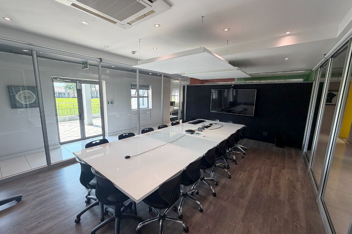 A modern, professional boardroom featuring glass partitions, laminate flooring, integrated ceiling air conditioning, and a wall-mounted display screen for presentations.
