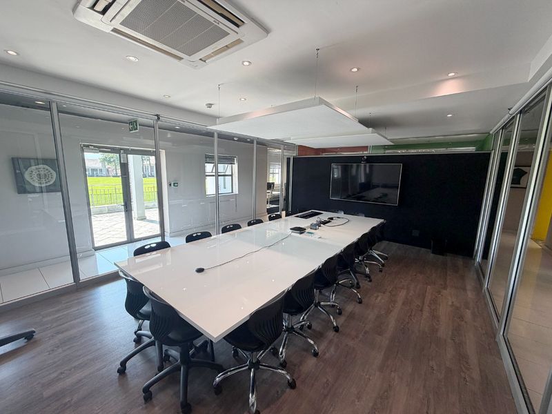 A modern, professional boardroom featuring glass partitions, laminate flooring, integrated ceiling air conditioning, and a wall-mounted display screen for presentations.