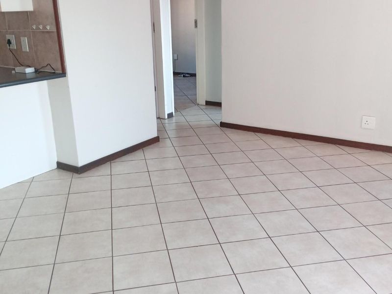 An open-plan living area featuring neutral ceramic floor tiles, clean white walls, and a partial view into the kitchen and hallway.