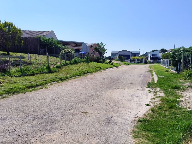 A view of an unpaved residential street with surrounding properties, wire fencing, and grass verges in a quiet neighborhood setting.
