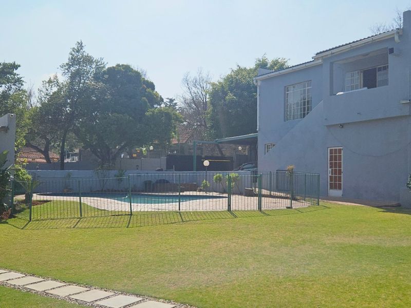 A spacious rear garden featuring a swimming pool with safety fencing, a detached garage, and a two-story house with a balcony.