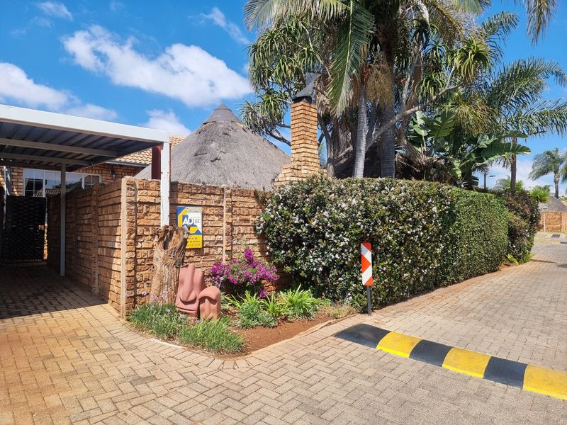 Front exterior view of the townhouse featuring a paved driveway, carport, brick boundary wall, and traditional thatched roof architecture.