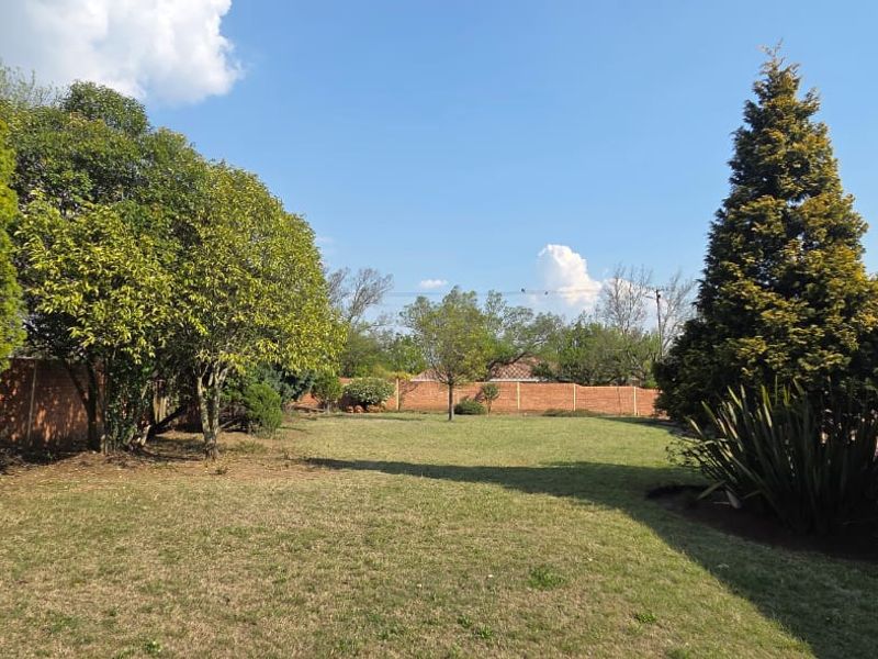 A spacious backyard garden featuring a well-maintained lawn, mature trees, and a brick boundary wall, adjacent to the main house structure.