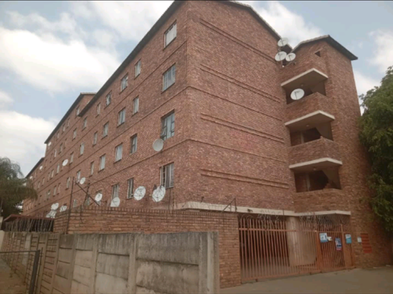 Exterior view of a multi-story brick apartment building featuring security fencing, gated access, and multiple satellite dish installations on the facade.