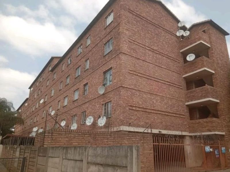Exterior view of a multi-story face brick apartment building featuring balconies, perimeter security fencing, and gated access.