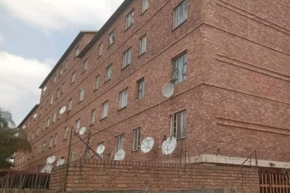 Exterior view of a multi-story face brick apartment building featuring security fencing and multiple satellite dish installations.