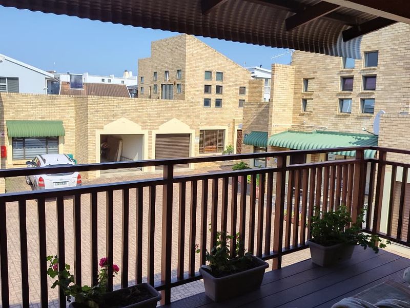 A covered balcony with timber decking and a wooden balustrade, overlooking a paved townhouse complex courtyard with face brick architecture.